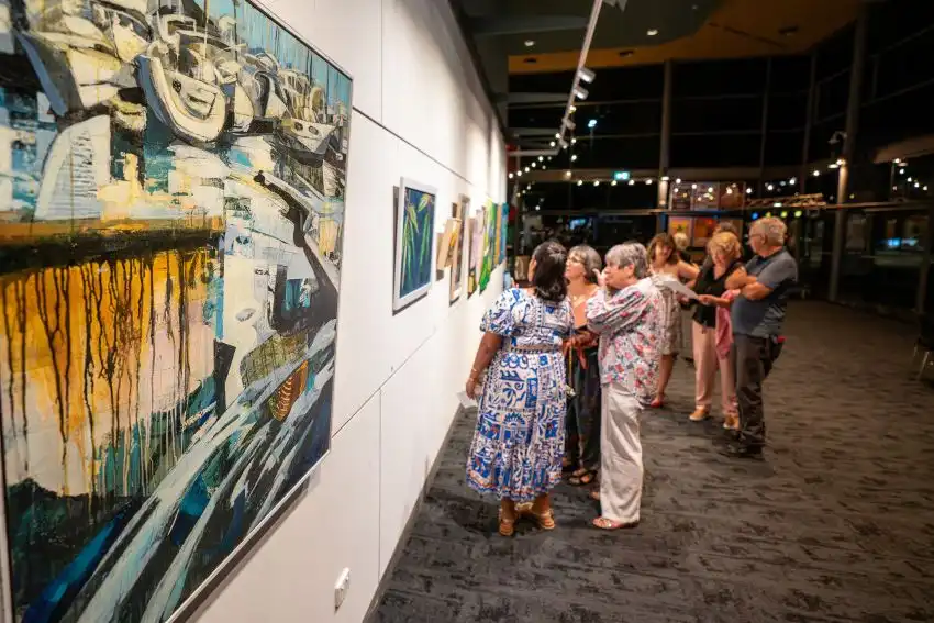 People viewing colourful artworks displayed along a gallery wall during an exhibition.
